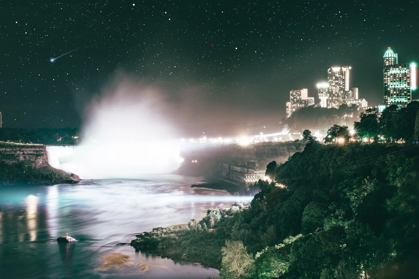 Tåge, der stiger op fra den oplyste kam af Niagara Falls over den mørke kløft, med byens lys og en stjerneklar himmel bagved