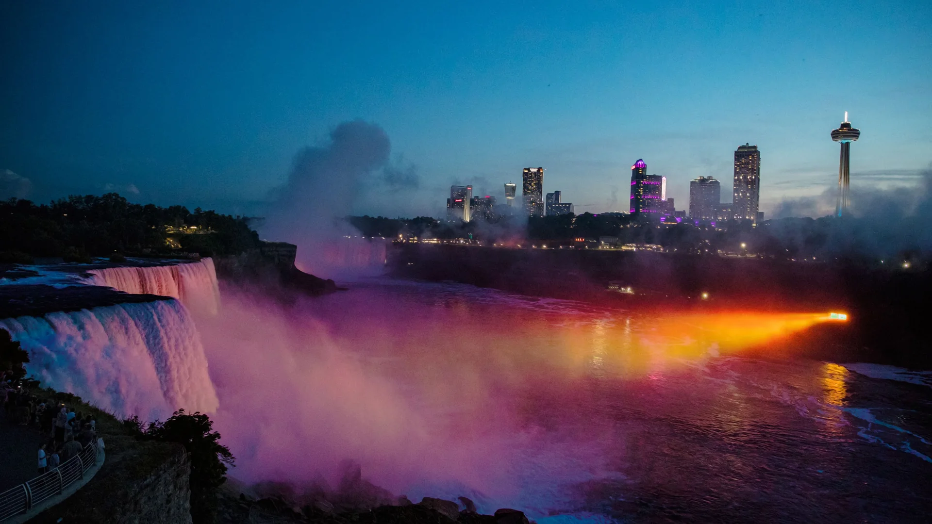 Dis og violet tåge stiger op fra foden af Niagara Falls ved skumringstid, med Skylon Tower og byens hoteltårne i baggrunden og en stråle af orange lys hen over floden