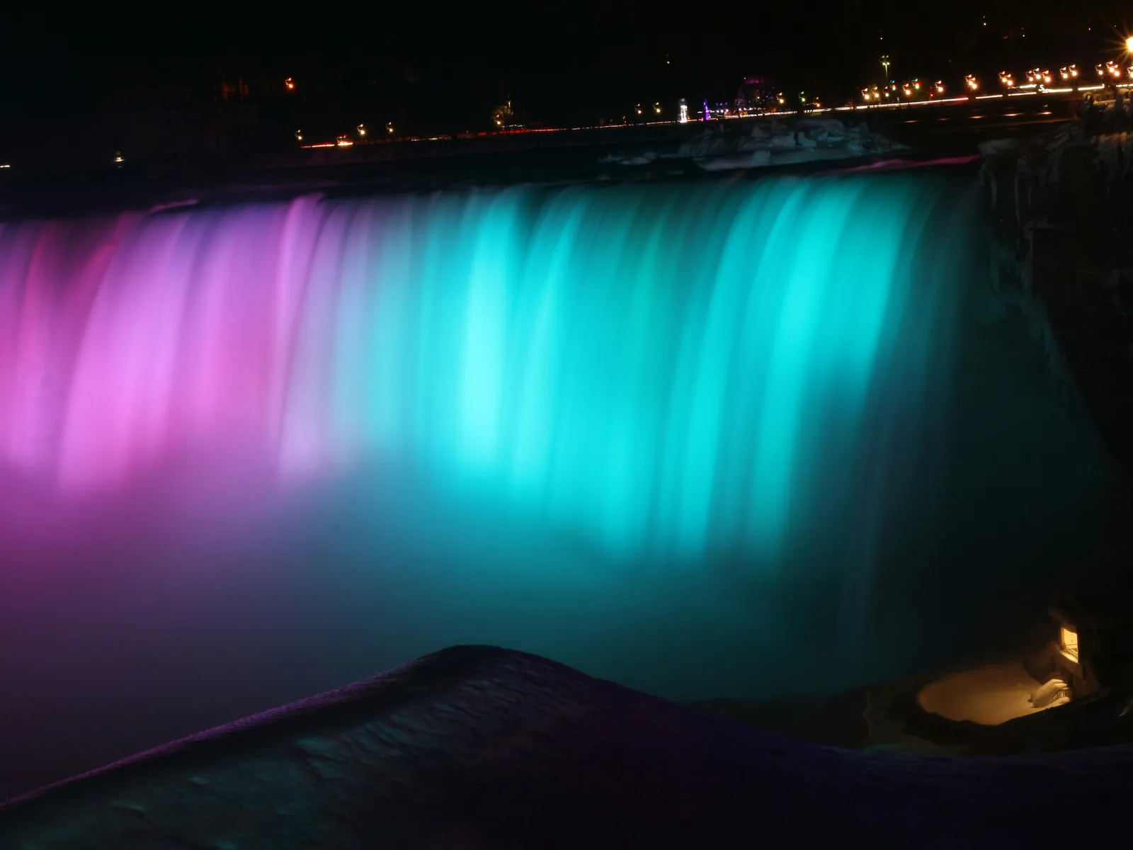 Niagara Falls oplyst i magenta og teal efter mørkets frembrud, set fra den canadiske side, med lysene fra vejen langs kløftens øverste kant